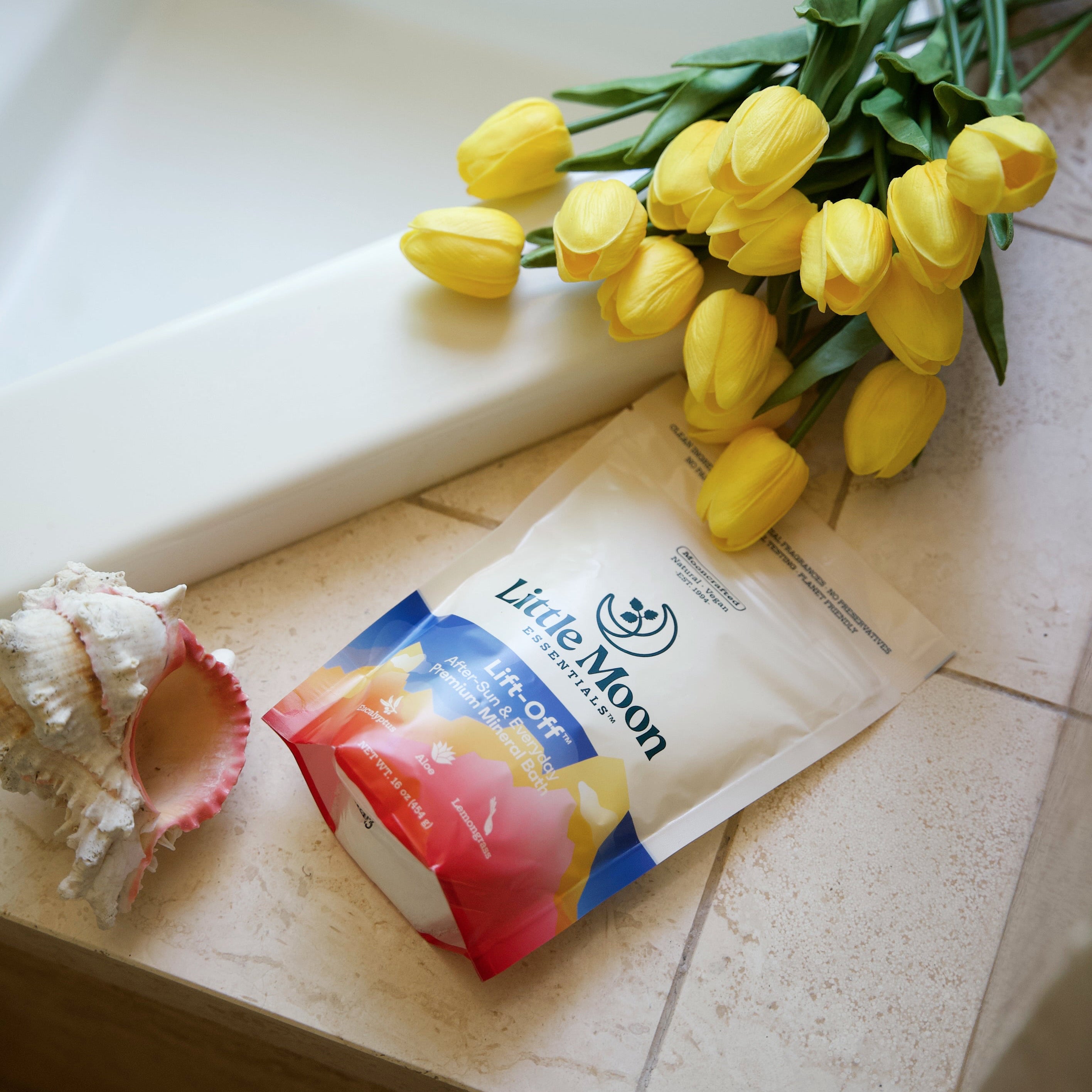A pouch of Little Moon Lift-Off Mineral Bath lies on a tiled bathroom counter next to a white bathtub, with yellow tulips and a seashell nearby.