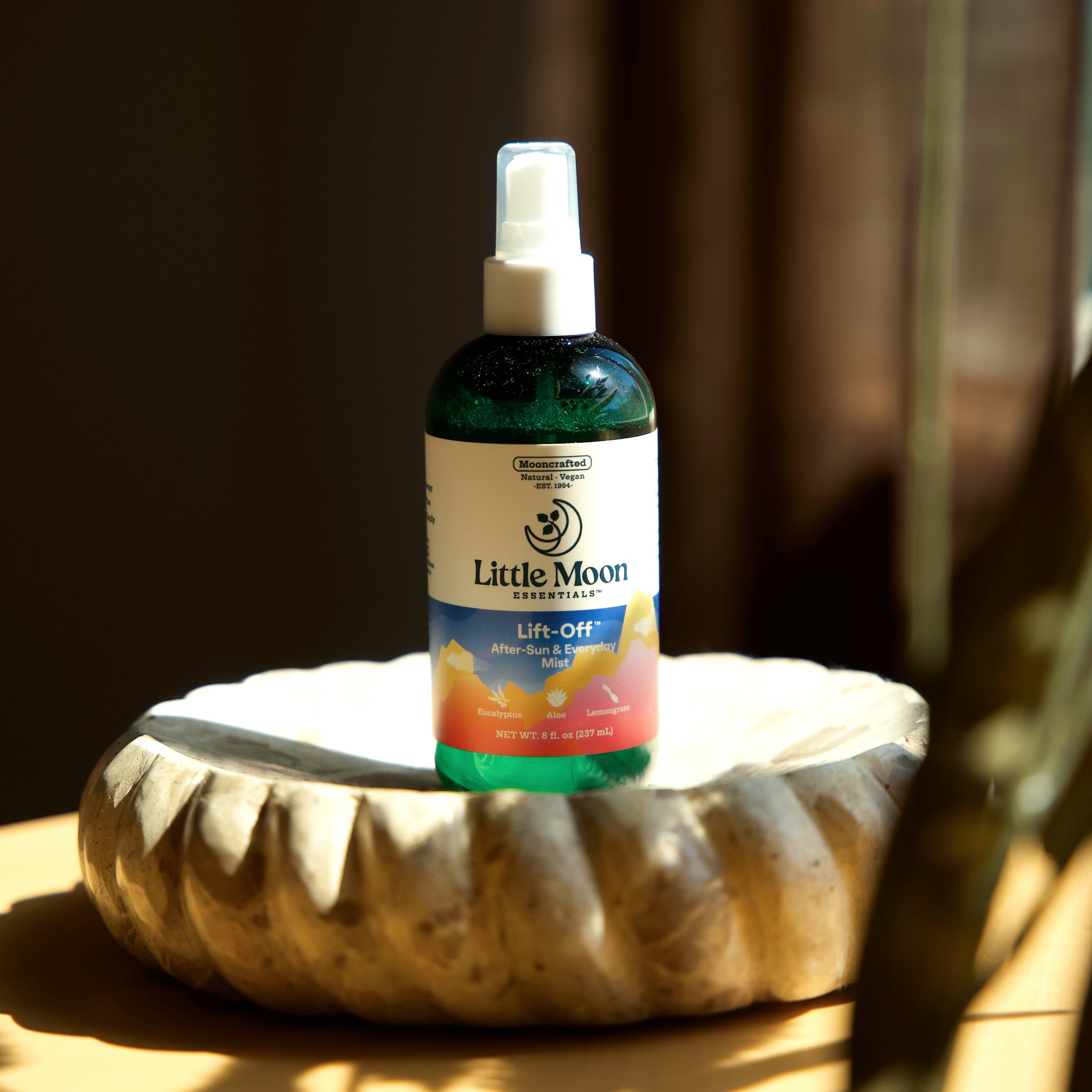 A bottle of Little Moon's Lift-Off After-Sun mist, featuring a white label and spray nozzle, resting on a textured ceramic dish against a dark brown background.