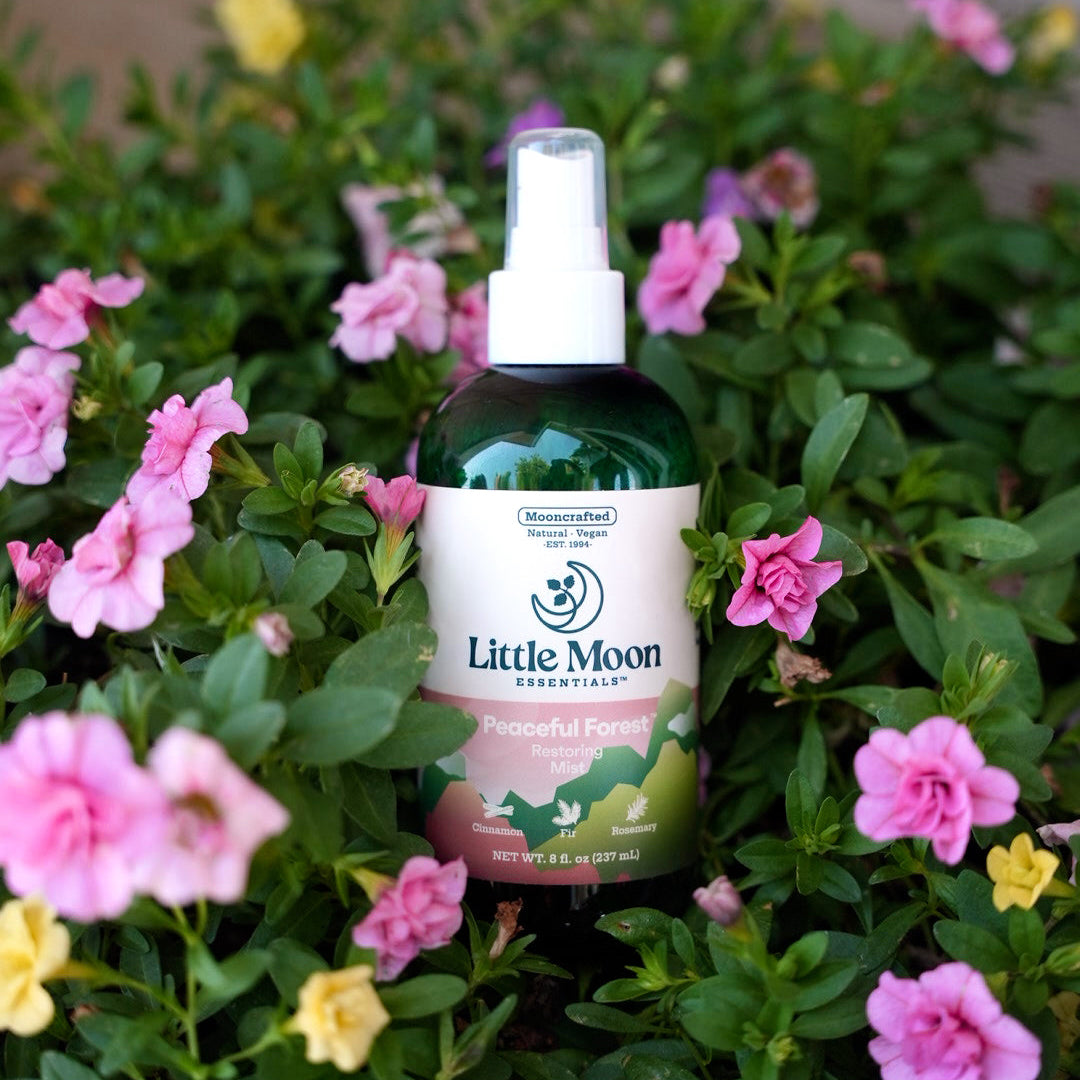 A bottle of Little Moon Natural Peaceful Forest spray nestled among green foliage and pink and yellow flowers.